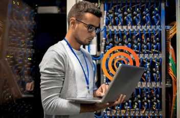 Young man with laptop standing by server cabinet while working with supercomputer